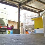Demon Town resident Tita Jorlang, at right in the background cooking outside on a fire that uses a fan’s cover as its grill, manages a family of eight from her small place in Majuro. Photo: Wilmer Joel.