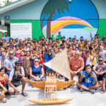 Rairok Elementary School students and teachers joined canoe program Director Alson Kelen, seated to right of outrigger canoe, after presentations at the school during Manit (Culture) Week. Photo: Chewy Lin.