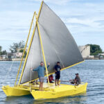 To support the sail, instead of a single mast, the yellow WAM catamaran has two poles positioned in an A-frame shape sitting on both hulls. The boat is steered by using a tiller arm that controls the two rudders, one on each hull.  The triangular sail sits in between the upper spar and lower boom, while the sail is controlled using a sheet (line), which passes from a fixed point through a pulley (or metal eye) and back to the person trimming (pulling or releasing depending on the direction of the wind and the desired course). The upper spar can be lowered to fold away the sail when it is not in use.   Crew and builders of the boat (not all of whom are pictured): Germany’s Henrik Richter-Alten (at bow), Isocker Anwel, Clancy Takia, Andy Caleb, Binton Daniel, Ken Daniel, George Lekjeb, Joe Lat.